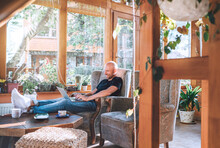 Man Dressed Black T-shirt And Jeans Sitting In A Comfortable Armchair, Using A Modern Slim Laptop And Drinking Tea In House Sunroom Living Room. Distance Or Freelance Or Writer Working Concept Image.