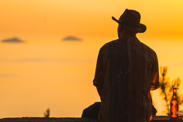 Back silhouette view of a man sitting on the wall and looking at beautiful sunset with clear sky in background