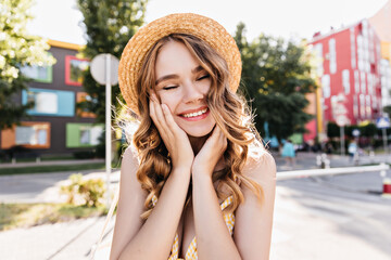 Wall Mural - Cute girl in hat laughing with eyes closed on city background. Romantic white woman dreamy posing in summer morning.