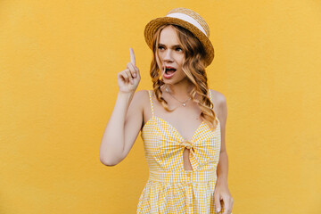 Wall Mural - Funny girl in stylish yellow dress posing in studio. Interested female model in straw hat looking around.