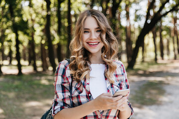 Wall Mural - Refined white woman in casual shirt waiting for call. Outdoor photo of relaxed girl holding smartphone on nature background.