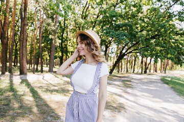 Wall Mural - Happy female model in cute summer attire posing in park. Outdoor shot of shy caucasian girl in hat.