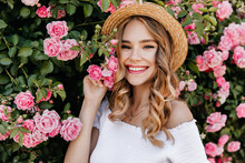 Adorable Girl With Curly Blonde Hair Posing In Garden. Portrait Of Caucasian Glad Woman Holding Pink Flower.