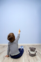 a child sitting on the floor with a bear cub points at a blue textured wall. copy space