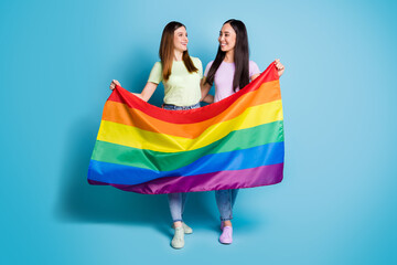 Poster - Full size photo of cheerful lesbians couple lovers walk foreign parade support tolerance same sex marriages carry gay rainbow flag wear t-shirts jeans shoes isolated blue color background