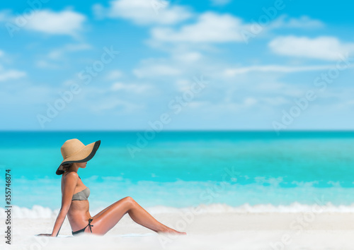Beach woman sun tanning sunbathing lying down on beach towel in sand body skincare sun exposure protection with hat for skin cancer.