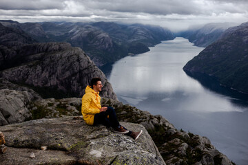 Wall Mural - Brunet man hiker in yellow raincoat smiles and sits on the cliff of Preikestolen mountain (Preacher's Pulpit or Pulpit Rock) with background of Lysefjord and low cloudy sky in Norway