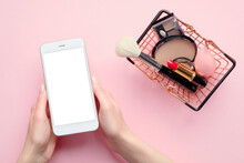 Buying Beauty Products Online Concept. Female Hand Holding Mobile Phone With Blank Screen Mockup And Shopping Cart With Cosmetics On Pink Background Top View.
