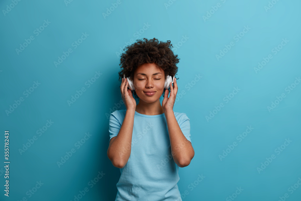 Satisfied curly young Afro American woman has carefree happy mood, closes eyes and listens music in headphones, wears casual blue t shirt, poses indoor. People, leisue, entertainment concept - obrazy, fototapety, plakaty 