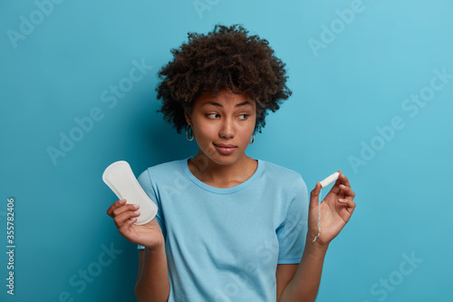 Fotomural Hesitant dark skinned woman holds unused clean sanitary napkin and tampon, chooses best intimate product for menstruation, cares about health, isolated on blue wall
