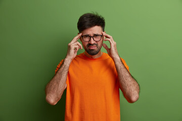 Wall Mural - Young man worker has unbearable headache, keeps hands on temples, frowns face from pain, feels painful migraine, overworked during preparing project, wears transparent glasses and orange t shirt