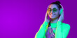 © Damir Khabirov - Horizontal banner of young smiling girl in neon green top and eyeglasses, listening to favorite tracks in headphones, isolated on purple background with copy space