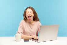 Naughty Overworked Businesswoman In Casual Clothes Showing Tongue Out, Expressing Disgust And Disobedience While Working On Laptop At Home Office. Indoor Studio Shot Isolated On Blue Background