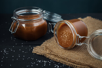 Two glass jars of homemade caramel with crystals of salt on black wooden table