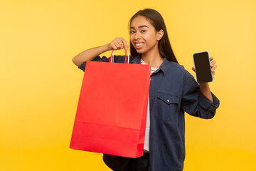 Portrait of happy shopper girl in stylish denim shirt holding bags and mobile phone with empty display mock up, satisfied with shopping in fashion store, thrift sale. indoor studio shot isolated