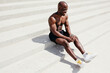 © DmitryStock - A black bare-chested fitness trainer sits and rests on the stairs in the park after intense outdoor exercise.