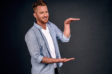 Horizontal shot of handsome Caucasian redhead man with bristle, shows height of something with both hands, pretends holding item or thing,demonstrates size, dressed in casual outfit, isolated on white