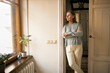 © shurkin_son - Indoor shot of serious sad middle aged female in casual clothes standing at doorway keeping arms crossed on her chest, looking through window, being upset while staying at home during quarantine