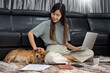 © snowing12 - Pretty asian woman working remotely from home using laptop sitting on the couch or sofa in living room for work online with pet puppy cute dog and guardian, work life balance concept.