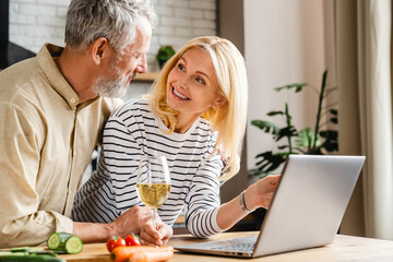 Caucasian middle aged couple watching show on laptop while cooking with wine at kitchen