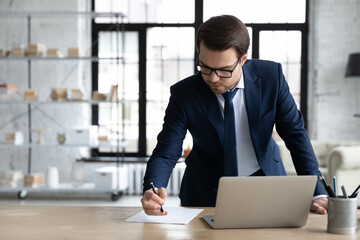 Serious Caucasian businessman in suit and glasses busy preparing business report or sign document in modern office, focused successful young male boss stand make notes working on laptop at workplace