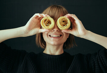 Portrait of a happy girl closes her eyes with nests of spaghetti macaroni with a smile on a black background.