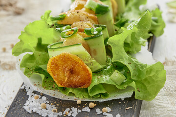 Sliced fried potatoes and cucumber on skewer on leaf of green lettuce on wooden Board. Sea salt is sprinkled in the foreground.