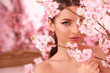 Closeup fashion spring face portrait of young beautiful caucasian woman with brunette hair in pony tail and perfect makeup looking through trees with pink flowers in blossom