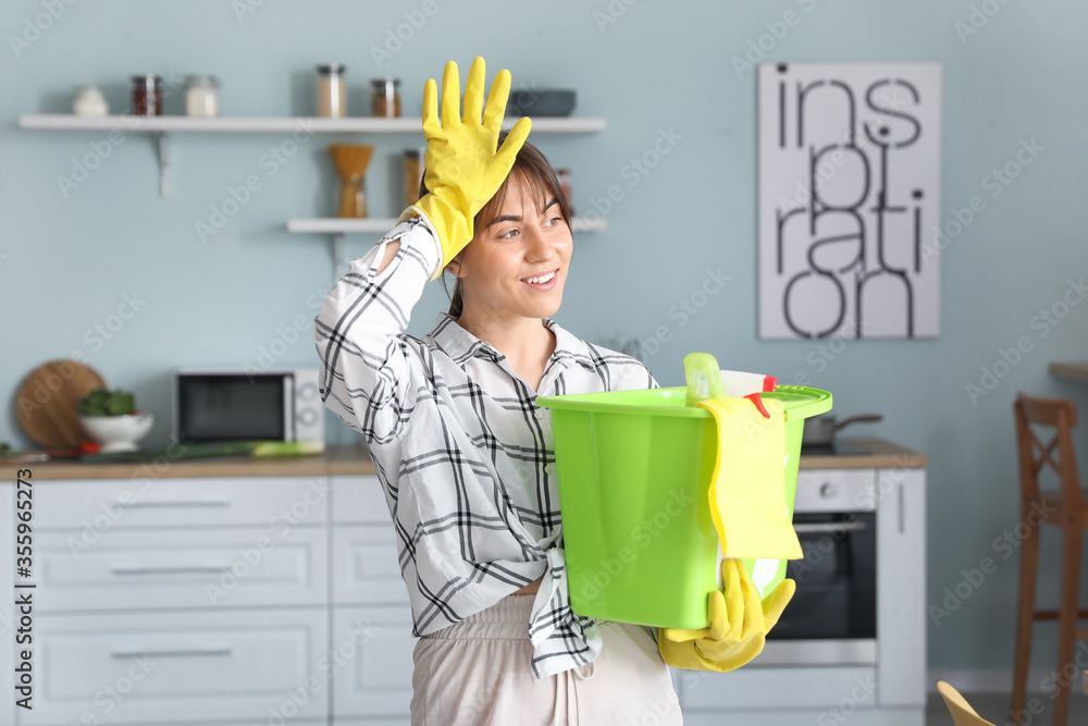 Tired young woman with cleaning supplies in kitchen