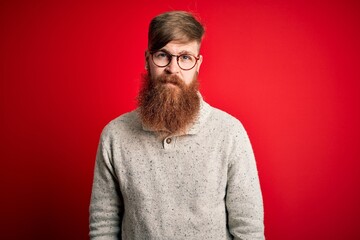 Canvas Print - Handsome Irish redhead man with beard wearing casual sweater and glasses over red background Relaxed with serious expression on face. Simple and natural looking at the camera.