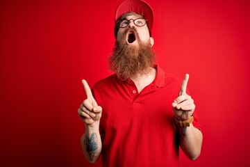 Sticker - Young handsome delivery man wearing glasses and red cap over isolated background amazed and surprised looking up and pointing with fingers and raised arms.