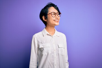 Wall Mural - Young beautiful asian girl wearing casual shirt and glasses standing over purple background looking away to side with smile on face, natural expression. Laughing confident.