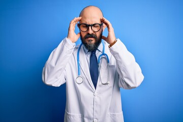 Canvas Print - Handsome bald doctor man with beard wearing glasses and stethoscope over blue background with hand on headache because stress. Suffering migraine.