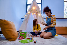 Caring For The Future. African American Woman Baby Sitter Entertaining Caucasian Cute Little Girl. They Are Playing With Dolls Together Sitting In Wigwam, Tent