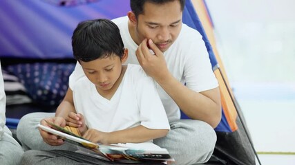 Sticker - Young man and his son reading story book together with camping tent background at home. Shot in 4k resolution