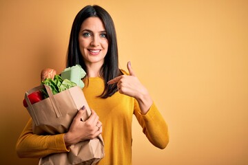 Wall Mural - Young brunette woman with blue eyes holding healthy groceries in paper bag from supermarket with surprise face pointing finger to himself