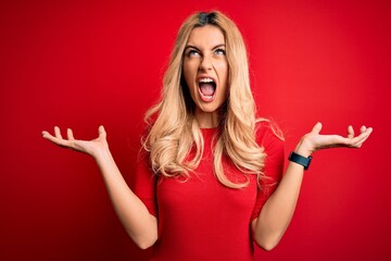 Poster - Young beautiful blonde woman wearing casual t-shirt standing over isolated red background crazy and mad shouting and yelling with aggressive expression and arms raised. Frustration concept.