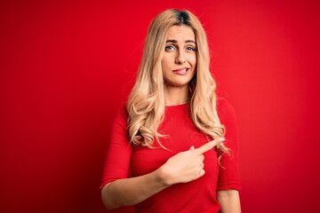 Wall Mural - Young beautiful blonde woman wearing casual t-shirt standing over isolated red background Pointing aside worried and nervous with forefinger, concerned and surprised expression