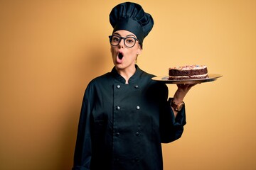 Canvas Print - Young beautiful brunette baker woman wearing cooker uniform and hat holding cake scared in shock with a surprise face, afraid and excited with fear expression