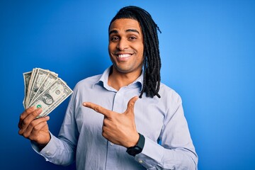 Wall Mural - Young african american afro man with dreadlocks holding dollars over blue background very happy pointing with hand and finger