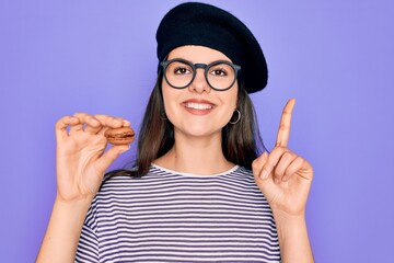 Sticker - Young beautiful girl wearing glasses and fashion beret holding french sweet pastry macaron surprised with an idea or question pointing finger with happy face, number one