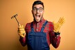 Young gardener man wearing working apron using gloves and tool over yellow background very happy and excited, winner expression celebrating victory screaming with big smile and raised hands