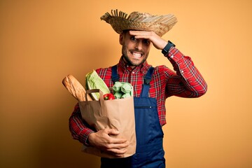 Sticker - Young rural farmer man holding fresh groceries from marketplace over yellow background stressed with hand on head, shocked with shame and surprise face, angry and frustrated. Fear and upset