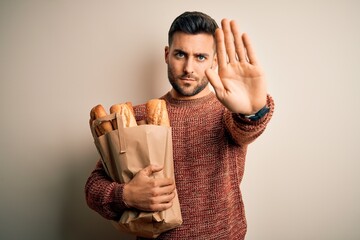 Wall Mural - Young handsome man holding groceries paper bag of fresh baguette bread over isolated background with open hand doing stop sign with serious and confident expression, defense gesture