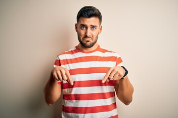 Canvas Print - Young handsome man wearing casual striped t-shirt standing over isolated white background Pointing down looking sad and upset, indicating direction with fingers, unhappy and depressed.