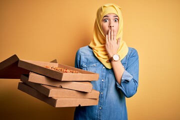 Poster - Young girl wearing muslim hijab holding boxes with Italian pizza over yellow background cover mouth with hand shocked with shame for mistake, expression of fear, scared in silence, secret concept