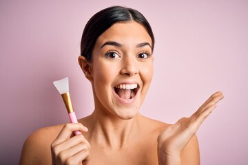 Poster - Young beautiful girl applying cosmetic using spatula over pink background very happy and excited, winner expression celebrating victory screaming with big smile and raised hands