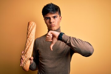 Poster - Young handsome man holding homemade fresh bread over isolated yellow background with angry face, negative sign showing dislike with thumbs down, rejection concept