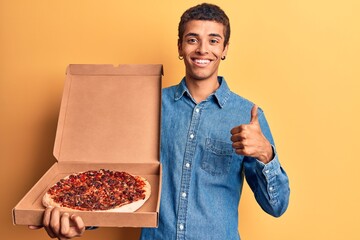 Sticker - Young african amercian man holding delivery pizza box smiling happy and positive, thumb up doing excellent and approval sign