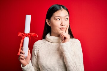 Canvas Print - Young asian student woman holding graduate diploma over red isolated background serious face thinking about question, very confused idea